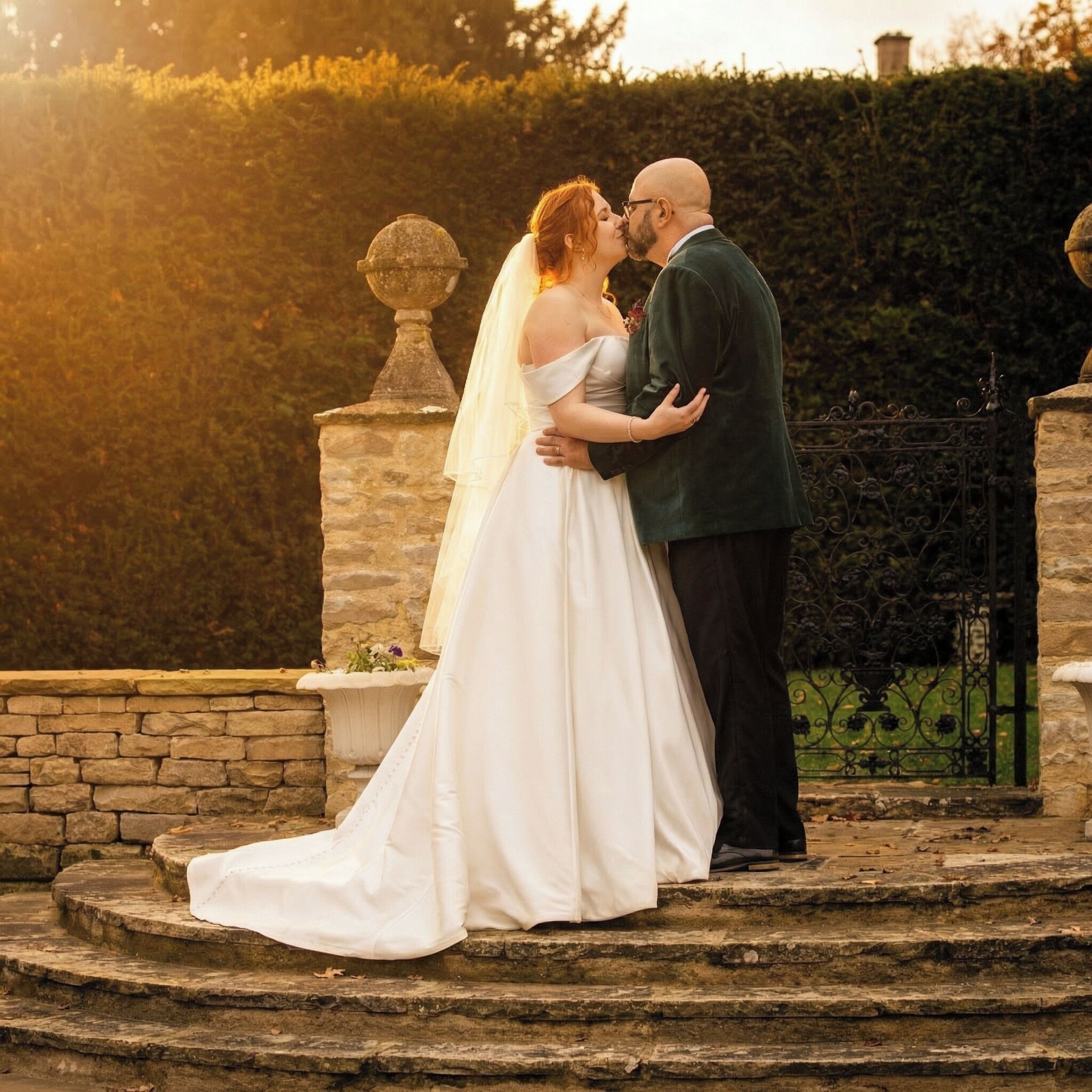 A couple embracing on stone steps with a stone balustrade and a hedge in the background.