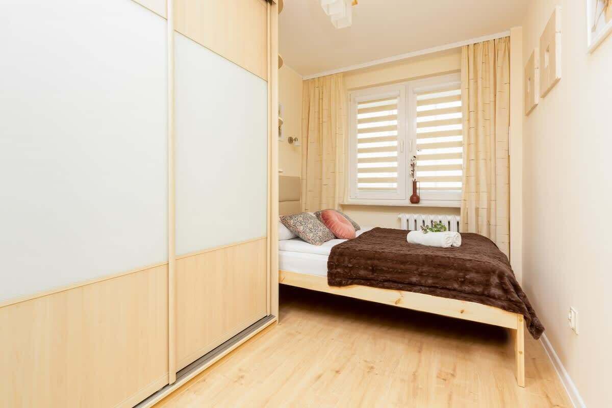 A small, well-lit bedroom with a wooden bed, a window with blinds, and a radiator.