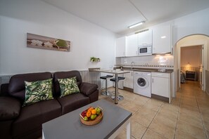A compact living space with a sofa, a small table with fruit, a washing machine, and a kitchenette. - Blue Coast (Haria)