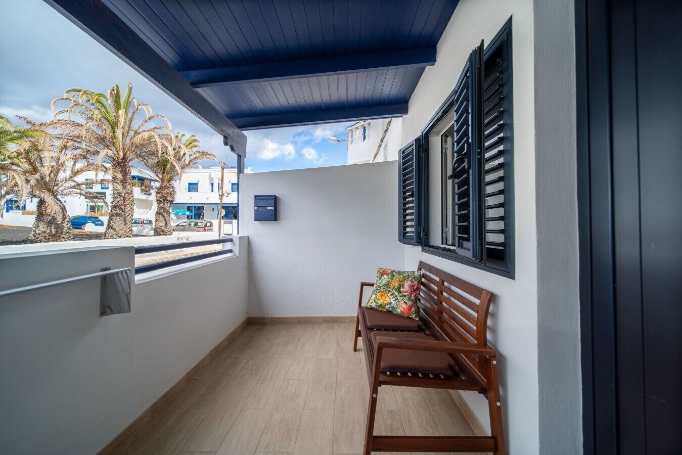 A balcony with a wooden bench, a blue ceiling, and a view of palm trees and buildings.