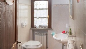 Ein kleines Badezimmer mit Holztür, Fenster mit Vorhängen, Toilette, Bidet, Waschbecken und Handtuchhalter.