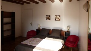 Une chambre avec un plafond en bois, un lit recouvert d’une couvre-lit à motifs floraux, deux tables de chevet avec des lampes, une chaise rouge et une bibliothèque.