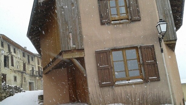 Un bâtiment à deux étages, doté de volets en bois, d’un escalier en pierre et d’un sol recouvert de neige.