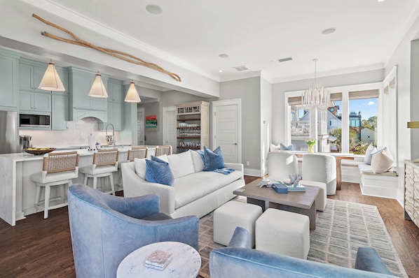 A modern living room with a white sofa, blue armchairs, and a coffee table. There is a kitchen area with light blue cabinets and a dining table with bar stools.
