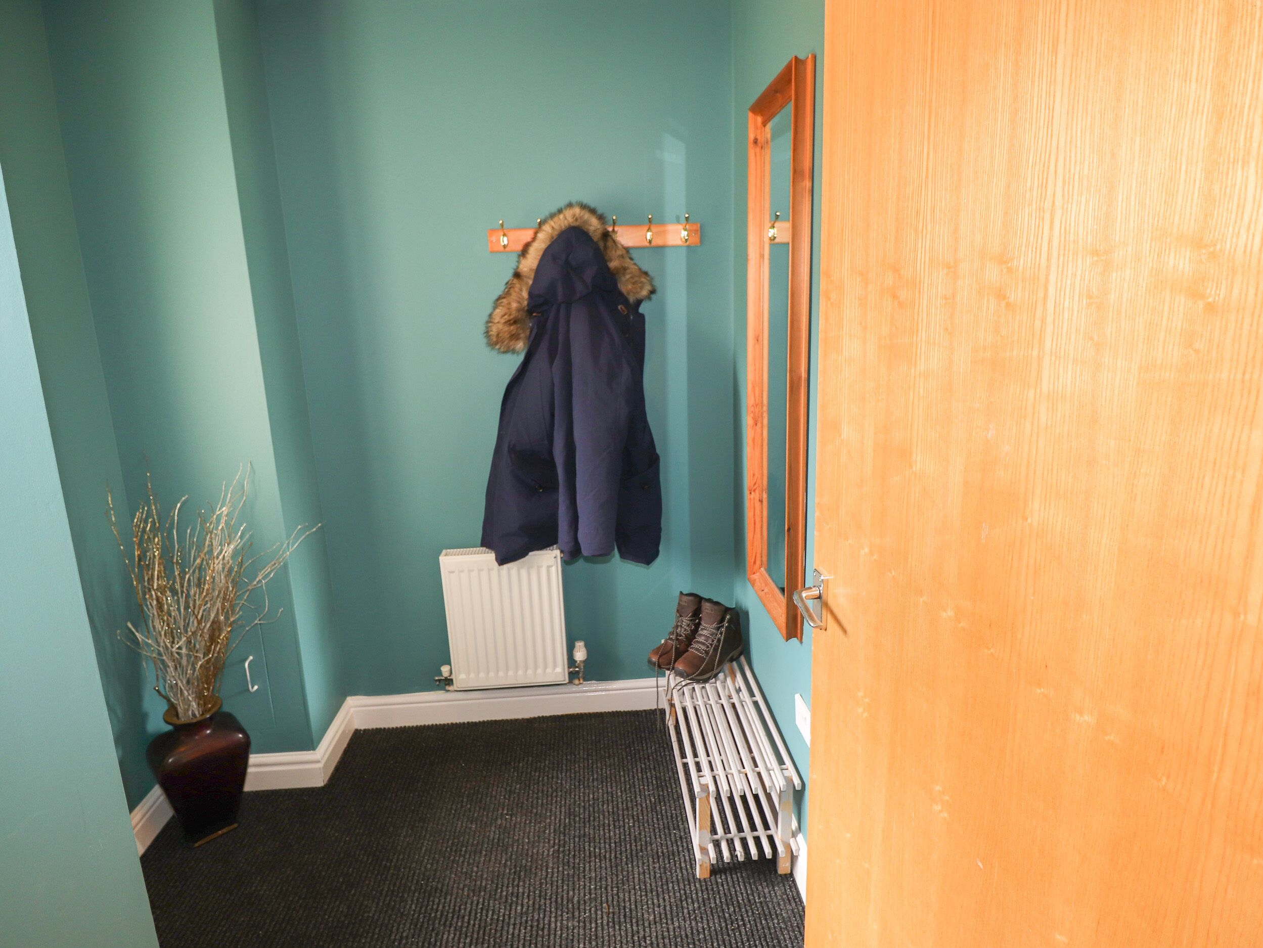 A hallway with teal walls, a radiator, a coat rack with a fur-lined coat, a wooden door, and a bench with shoes.