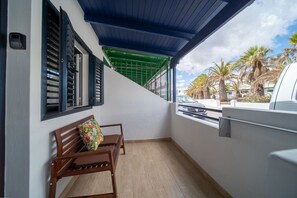 A balcony with a wooden bench, a green awning, and a view of palm trees and parked cars. - Green Coast (Punta Mujeres)