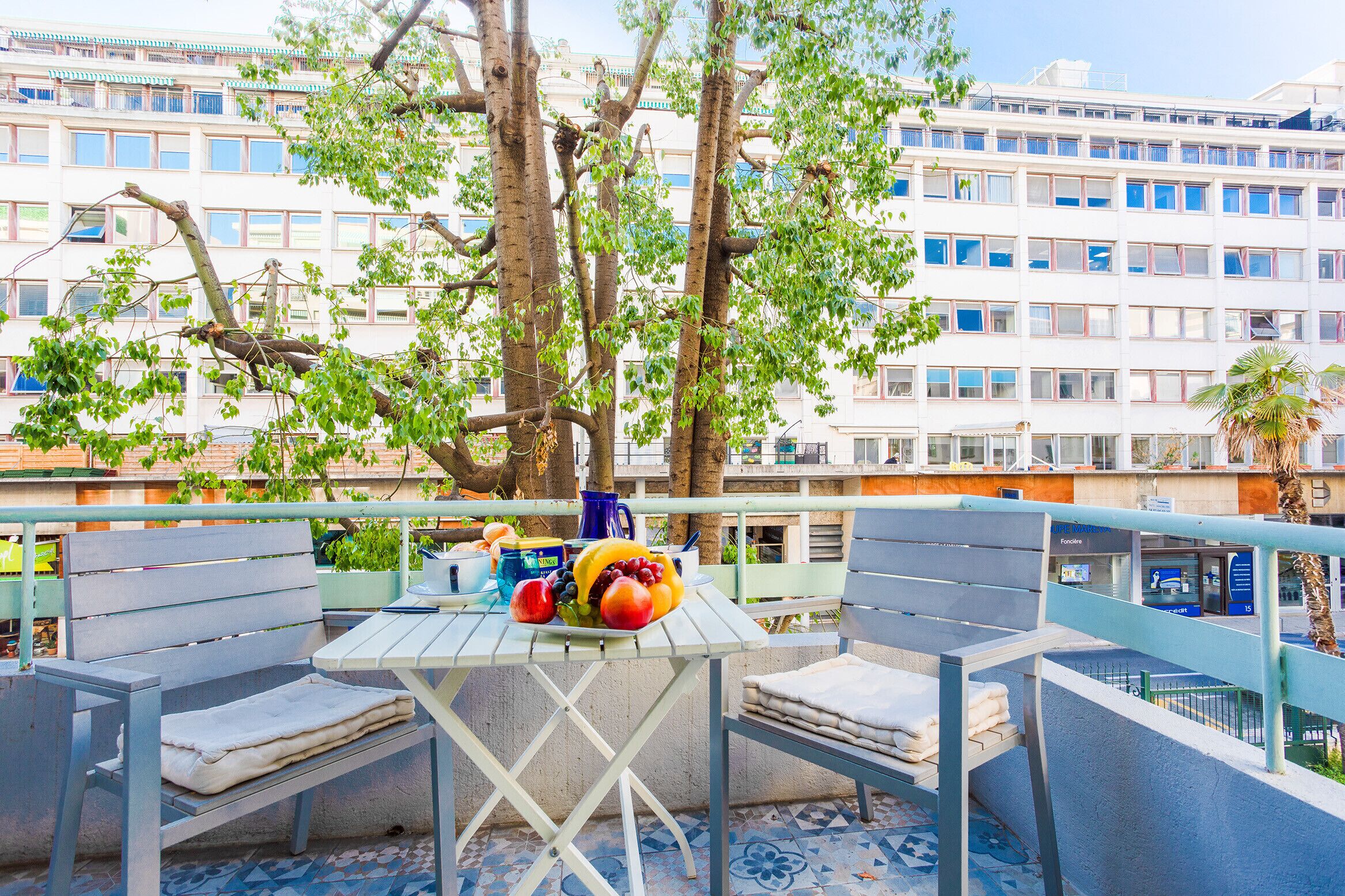 Een terras met een tafel voor twee personen, omringd door stoelen, met uitzicht op een gebouw met meerdere ramen en balkons.