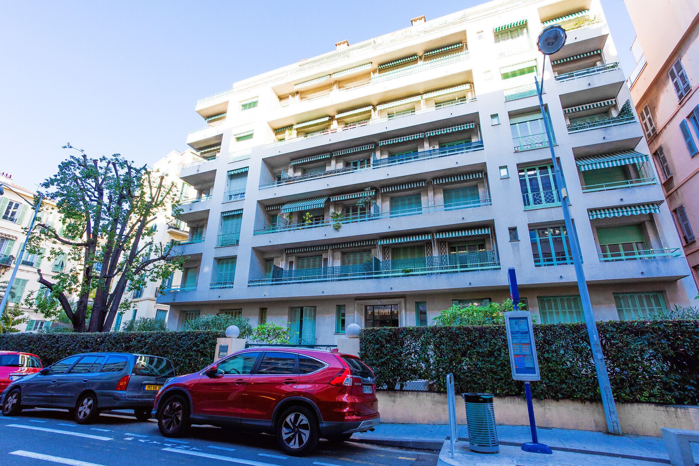 Um edifício com várias andares e varandas, uma rua com carros estacionados e um céu claro.