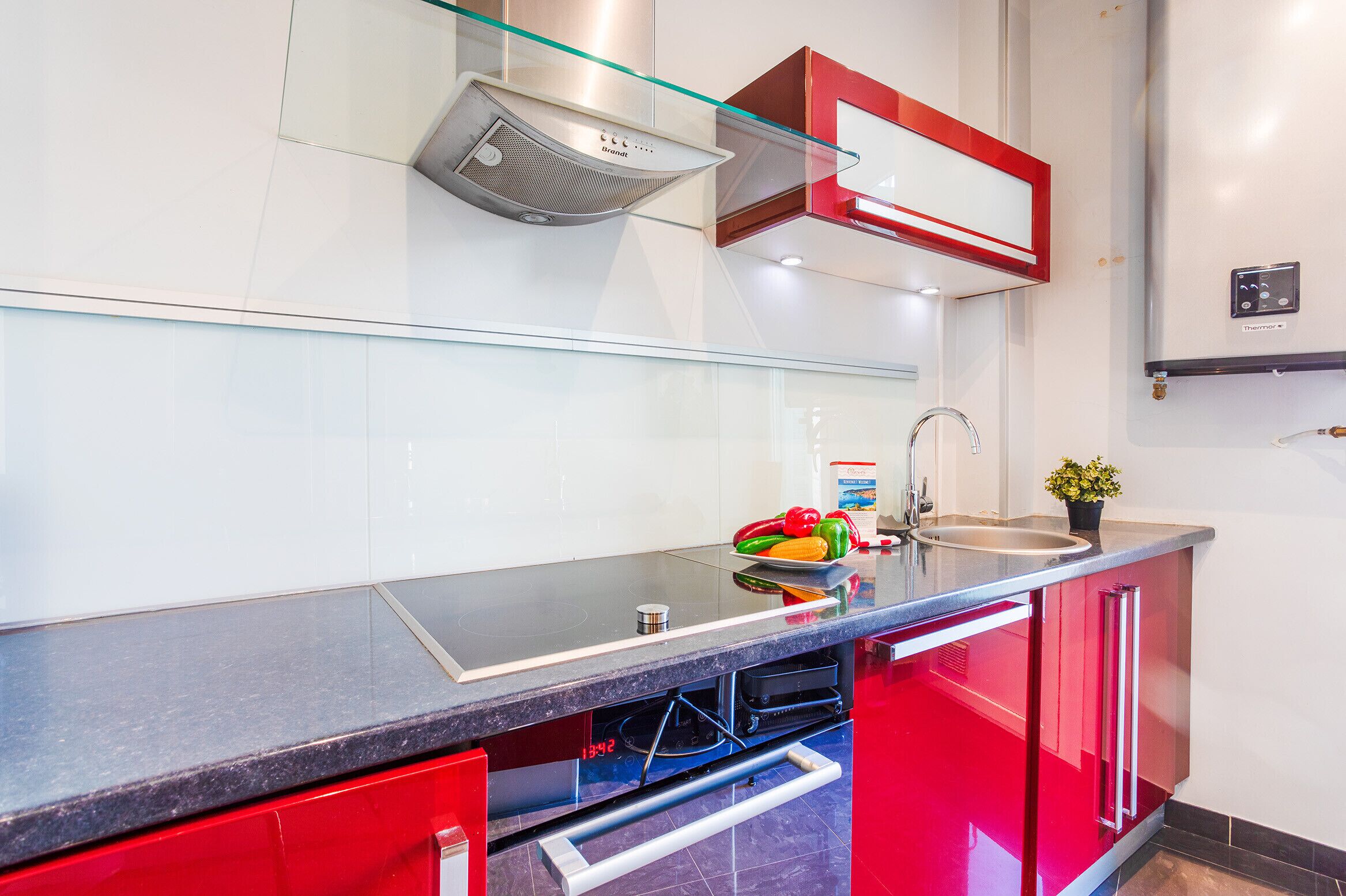 A modern kitchen with a glass-top countertop, red cabinets, a stainless steel range hood, and a built-in sink.