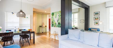A modern living room with a white sofa, a dining area with a table and chairs, and a kitchen in the background.
