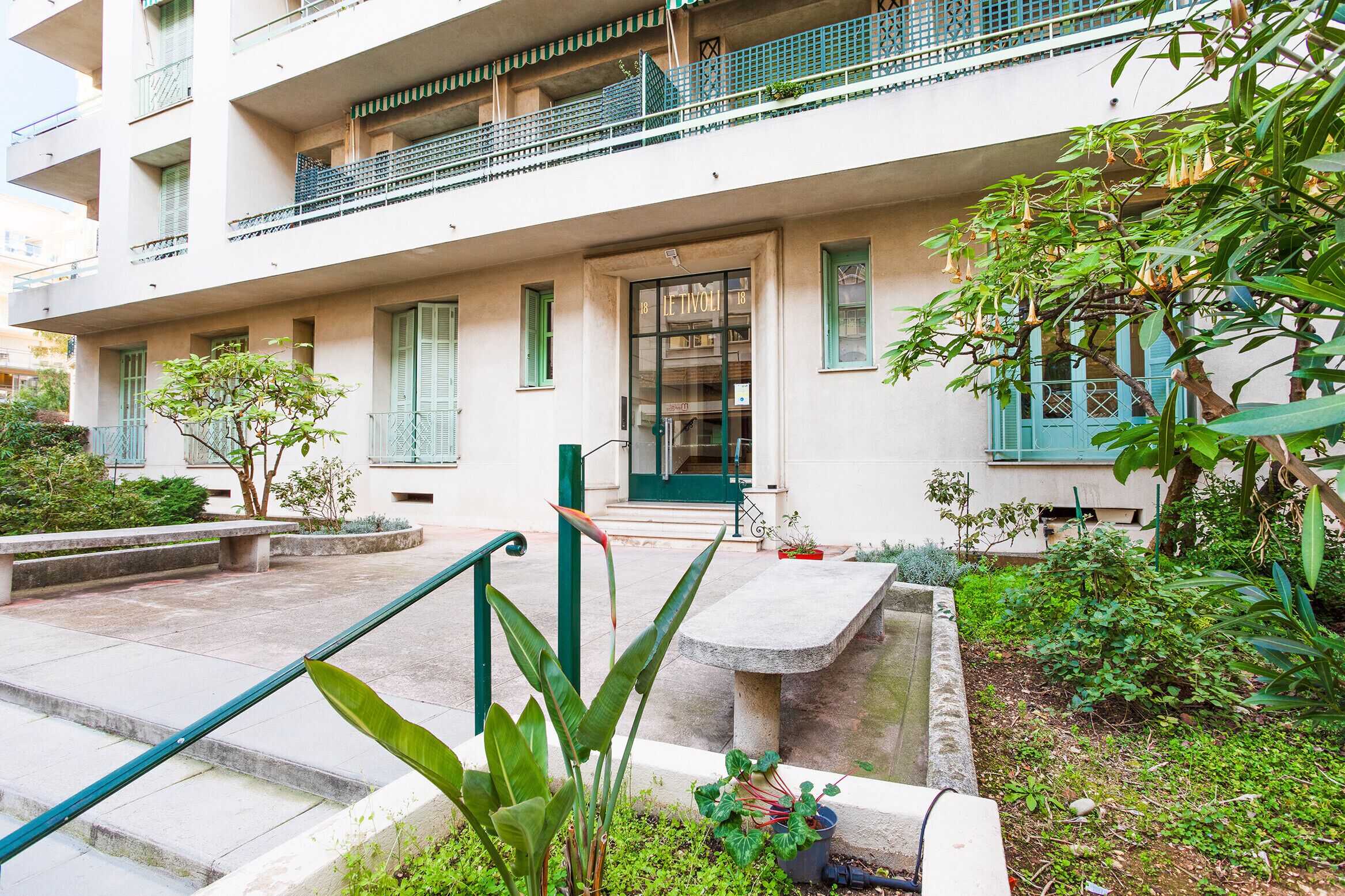 A building with a balcony, a door labeled "Le Tournai," and a small garden area with benches and potted plants.