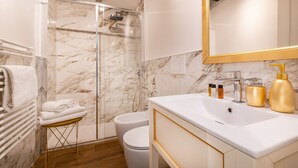 Une salle de bain moderne avec une douche à parois de verre, un lavabo blanc doté de robinetterie dorée et des murs en marbre.