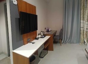 A compact hotel room with a flat-screen TV mounted on a wooden panel, a small desk with a telephone, a coffee maker, and a chair. - RG Exclusive Stay & Fine Dine, Akola (Akola)