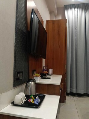 A hotel room with a flat-screen TV mounted on a wall, a small desk with a kettle, cups, and a tray of snacks, a wooden wardrobe, and a window with curtains.