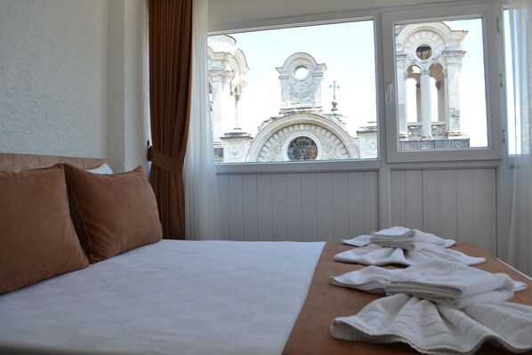 Una habitación de hotel con una cama, almohadas marrones, y una ventana que ofrece vistas a un edificio con columnas y un reloj.