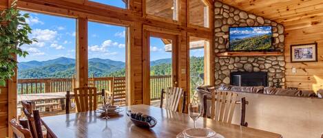 A wooden cabin with a dining area, stone fireplace, and a scenic mountain view.