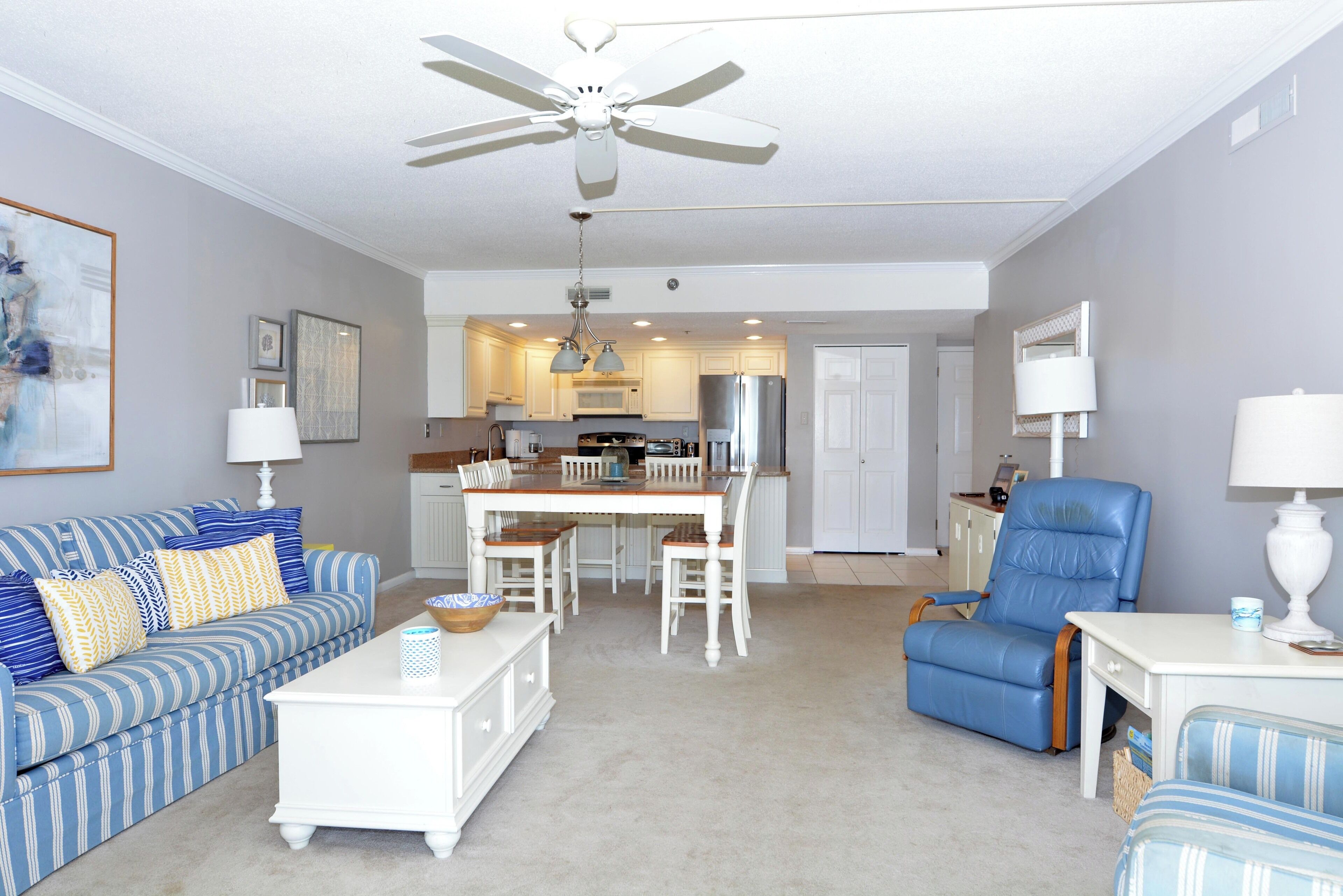A living room with a sofa, coffee table, and a dining table with chairs. There is a ceiling fan, kitchen area with cabinets and appliances, and a blue armchair.