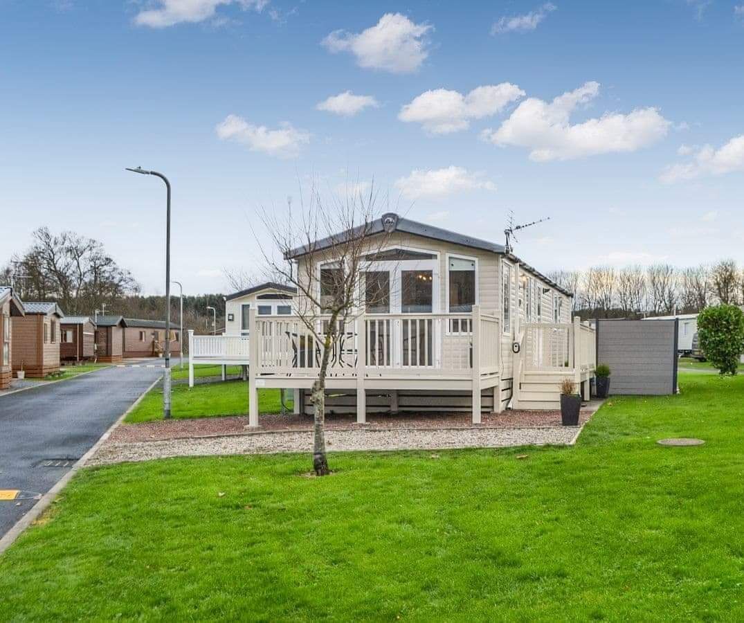 A mobile home with a porch, a small deck, and a shed in a green, grassy area.