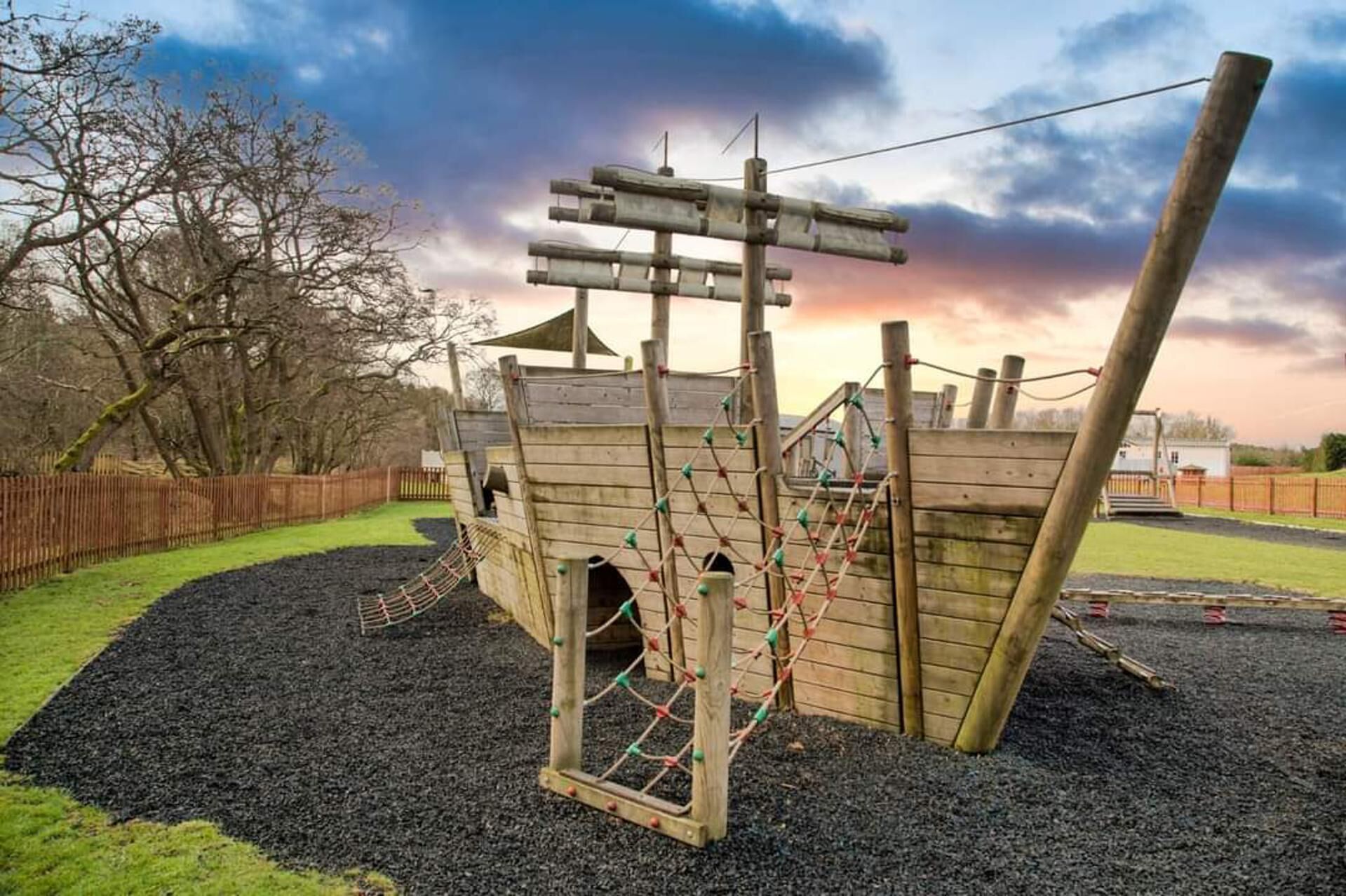 A wooden play structure with climbing nets and a slide, surrounded by a fence and trees.