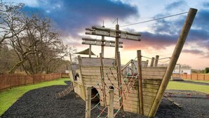 A wooden play structure with climbing nets and a slide, surrounded by a fence and trees. - Apple Tree Lodge, Hot Tub, 3 Bedroom, 2 Bars (Morpeth)