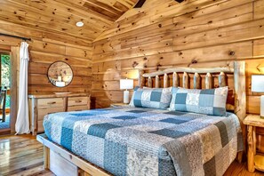 A wooden cabin bedroom with a bed, a round mirror, a nightstand, and a window with a curtain. - Riverfront, ½ Mile to Smoky Mtn National Park (Townsend)