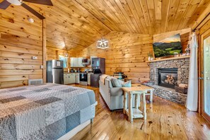 A log cabin interior with a bed, sofa, dining table, kitchen, fireplace, and a mounted TV. - Riverfront, ½ Mile to Smoky Mtn National Park (Townsend)