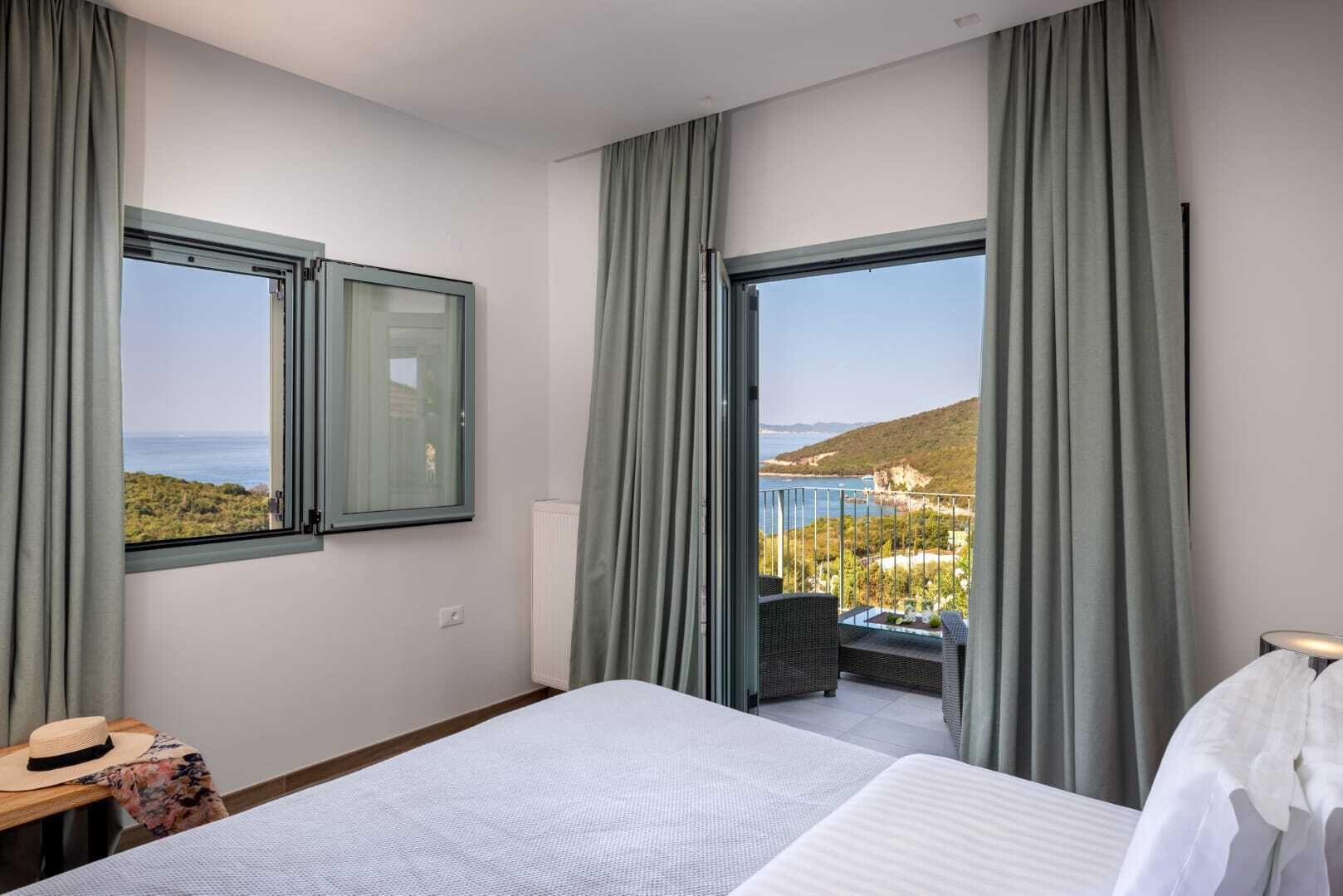 Une chambre d’hôtel avec un grand lit, deux fenêtres avec des rideaux, une vue sur la mer et un balcon équipé de mobilier d’extérieur.