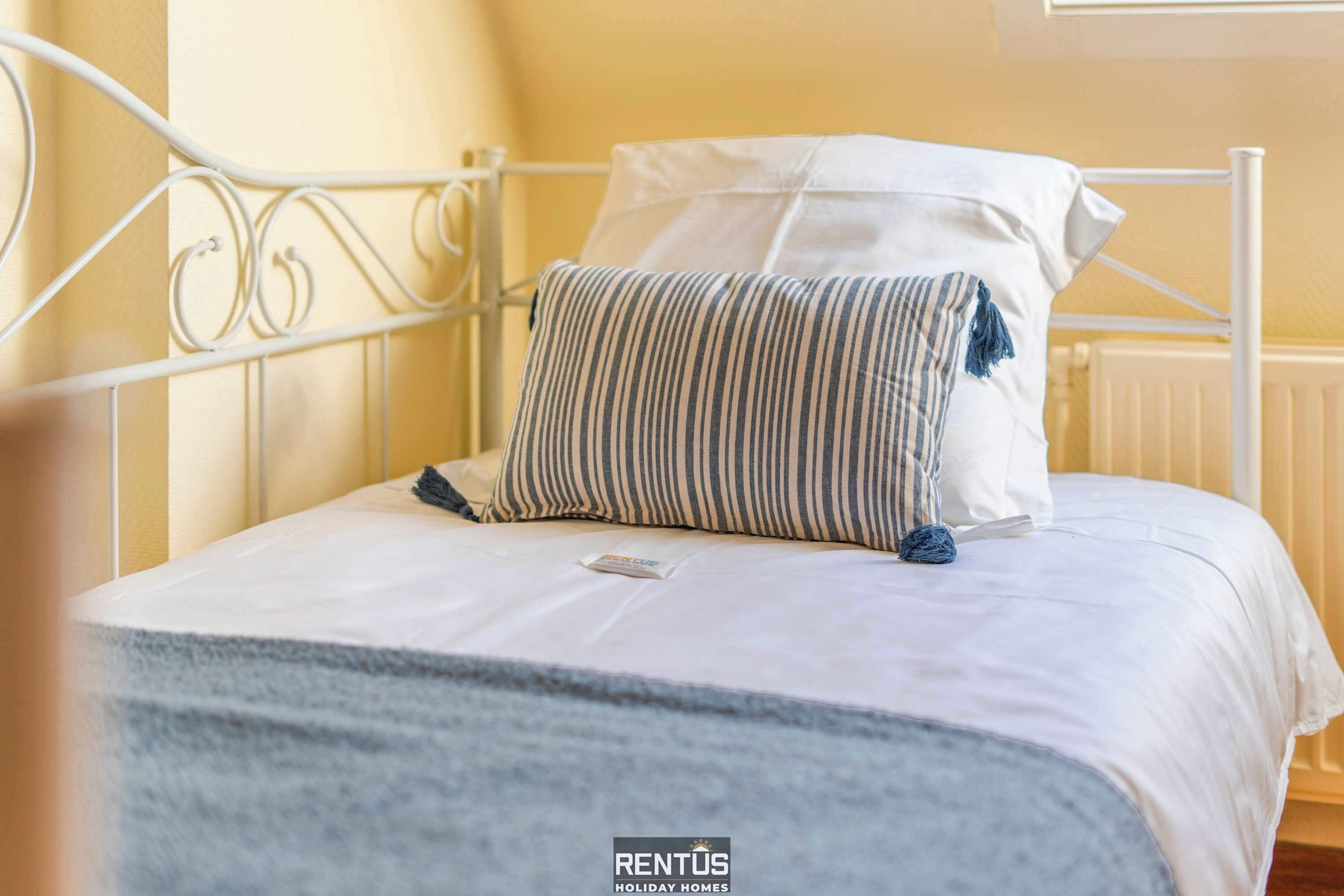 A neatly made bed with a striped pillow and a white pillow, a white metal headboard, and a window with blinds.