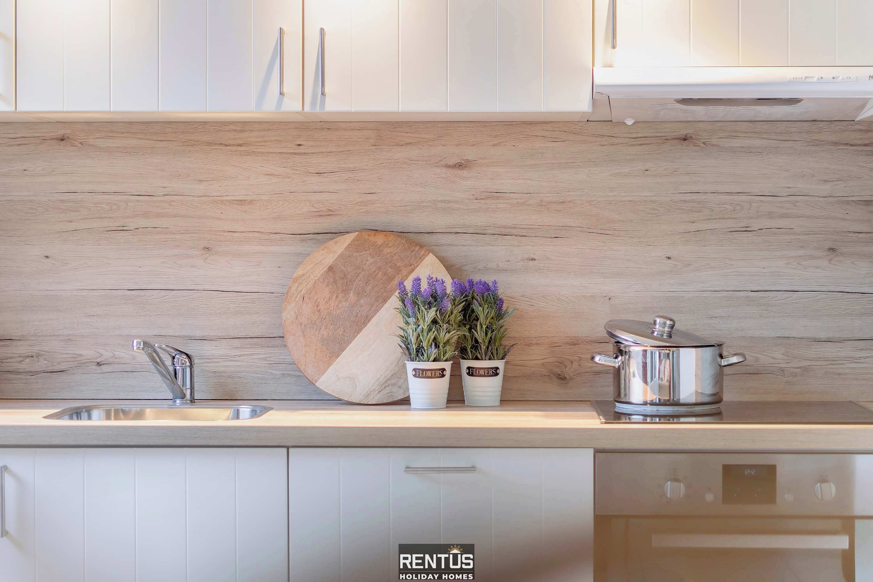A modern kitchen with white cabinets, a wooden backsplash, a sink, two potted plants, and a stainless steel pot.
