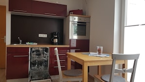 A compact kitchen with a dishwasher, a coffee maker, and a small oven. There is a wooden table with two chairs and a glass of water. - House (Dornum)