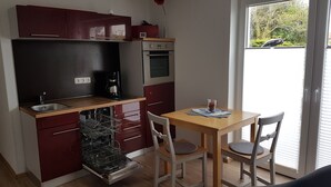 A compact kitchen with a dishwasher, a coffee maker, and a small oven. There is a dining table with two chairs and a window with blinds. - House (Dornum)