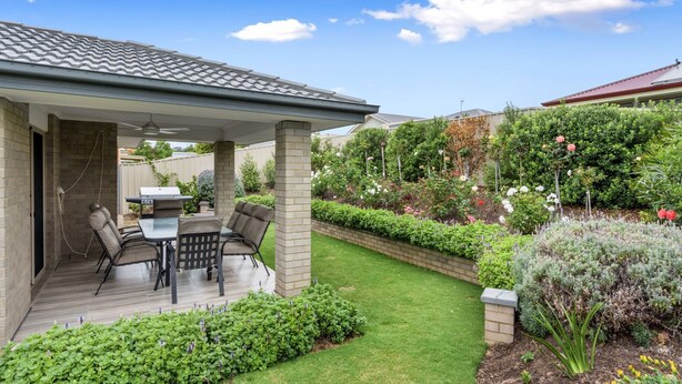 Een overdekt terras met een barbecue, buitenmeubilair en een keurig onderhouden tuin met diverse planten en bloemen.