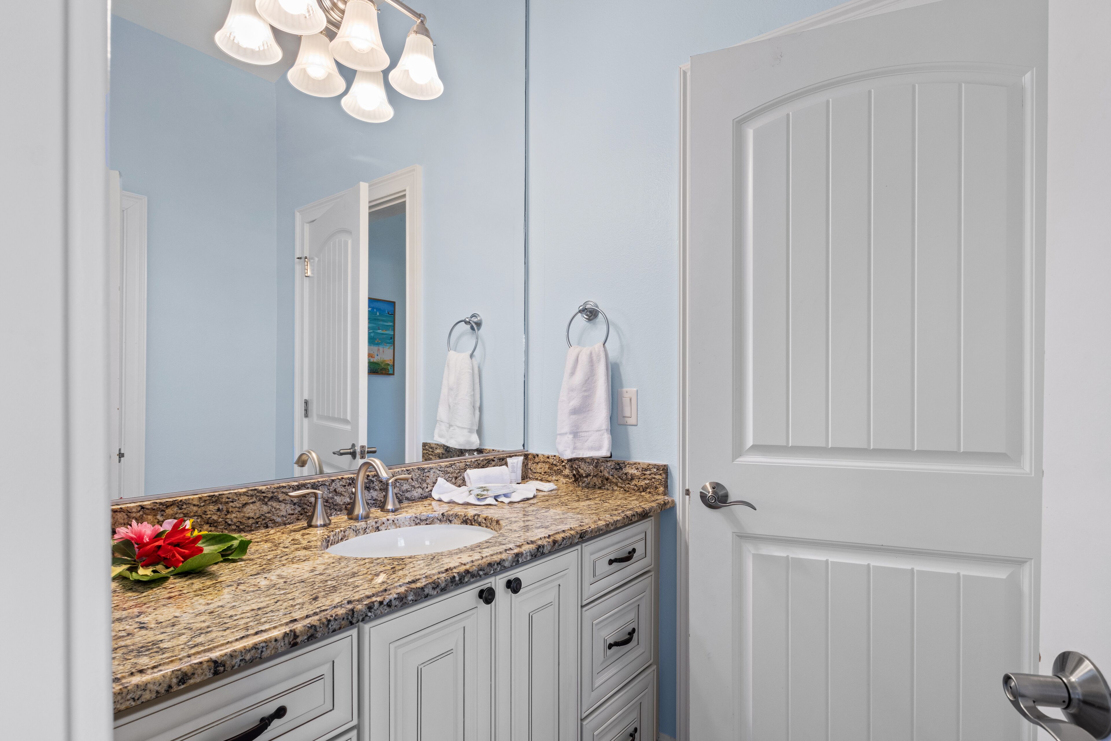 A bathroom with a granite countertop, white cabinets, a sink, and a mirror. There are two towels hanging on hooks and a flower arrangement on the countertop.