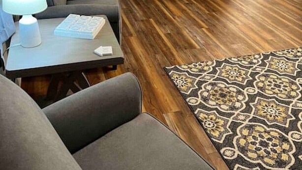 Un salon avec un téléviseur à écran plat fixé au mur, un canapé gris, une petite table avec une lampe et un tapis à motifs.