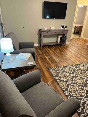 Ein Wohnzimmer mit flachbildfernseher an der Wand, einem grauen Sofa, einem kleinen Tisch mit Lampe und einem gemusterten Teppich.