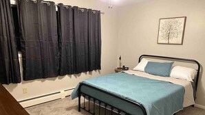 Une chambre à coucher avec un lit, une table de chevet, un radiateur et une fenêtre avec des rideaux.
