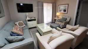 Una sala de estar con un sofá, dos sillones, una mesa de centro, un televisor, una lámpara y un cuadro en la pared.