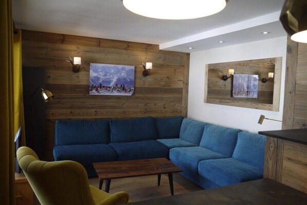 A modern living room with a blue sectional sofa, a yellow armchair, a wooden coffee table, and a wooden accent wall with a framed picture.