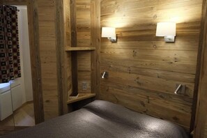 A room with wooden panel walls, a bed with a textured blanket, two wall-mounted light fixtures, and a small shelf with a book.