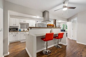 Una cocina moderna con armarios blancos, una isla central con taburetes rojos, una campana extractora de acero inoxidable y un ventilador de techo.