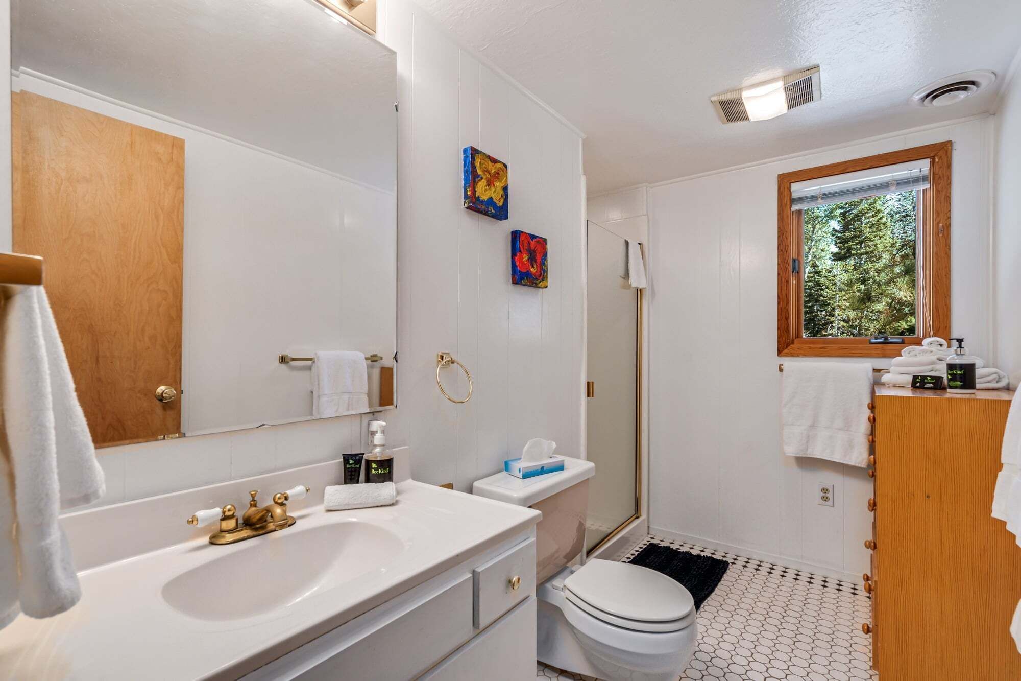 Un baño con un espejo grande, un lavabo, un inodoro y una ducha. Hay una ventana con vistas a árboles y una puerta de madera.
