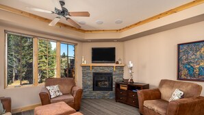 A cozy living room with a fireplace, comfortable seating, and a large window offering a view of the forest. - Modern Condo- Jacuzzi Tub SM #201 by Bear Valley Vacation Rentals (Arnold)