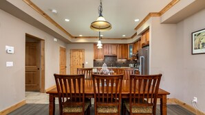 A dining area with a wooden table and chairs, a kitchen in the background, and a ceiling with recessed lighting. - Modern Condo- Jacuzzi Tub SM #201 by Bear Valley Vacation Rentals (Arnold)