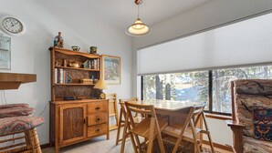 A cozy room with a wooden dining table, chairs, a bookshelf, and a window with a view of trees. - Year Round Condo- Sleeps 5! - Tamarack #17 by Bear Valley Vacation Rentals (Arnold)