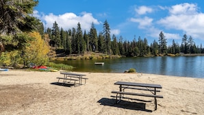 A sandy beach with two picnic tables and a lake surrounded by trees. - Year Round Condo- Sleeps 5! - Tamarack #17 by Bear Valley Vacation Rentals (Arnold)