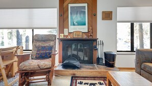 A cozy living room with a fireplace, a large window, and a wooden chair with a patterned cushion. - Year Round Condo- Sleeps 5! - Tamarack #17 by Bear Valley Vacation Rentals (Arnold)