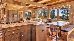 A rustic cabin interior with a wooden dining table, bar stools, a stone wall, and large windows offering a forest view. - Overlooking Beautiful Bear Lake VH#318 by Bear Valley Vacation Rentals (Arnold)