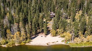 An aerial view of a cabin surrounded by dense forest, a sandy beach, and a small dock. - Overlooking Beautiful Bear Lake VH#318 by Bear Valley Vacation Rentals (Arnold)