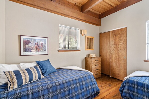 Un dormitorio con dos camas, vigas de madera en el techo, un cuadro enmarcado en la pared, una ventana con persianas y un armario de madera.