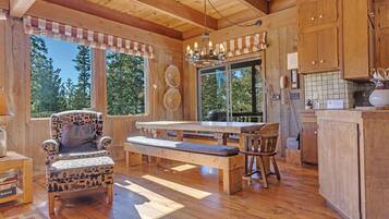 A wooden cabin interior with a dining table, benches, a patterned armchair, and a kitchen area with wooden cabinets and a hanging telephone.